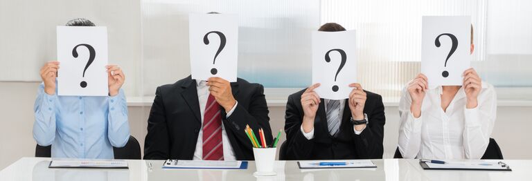 Four people sitting at a table, holding signs with question-marks over their faces